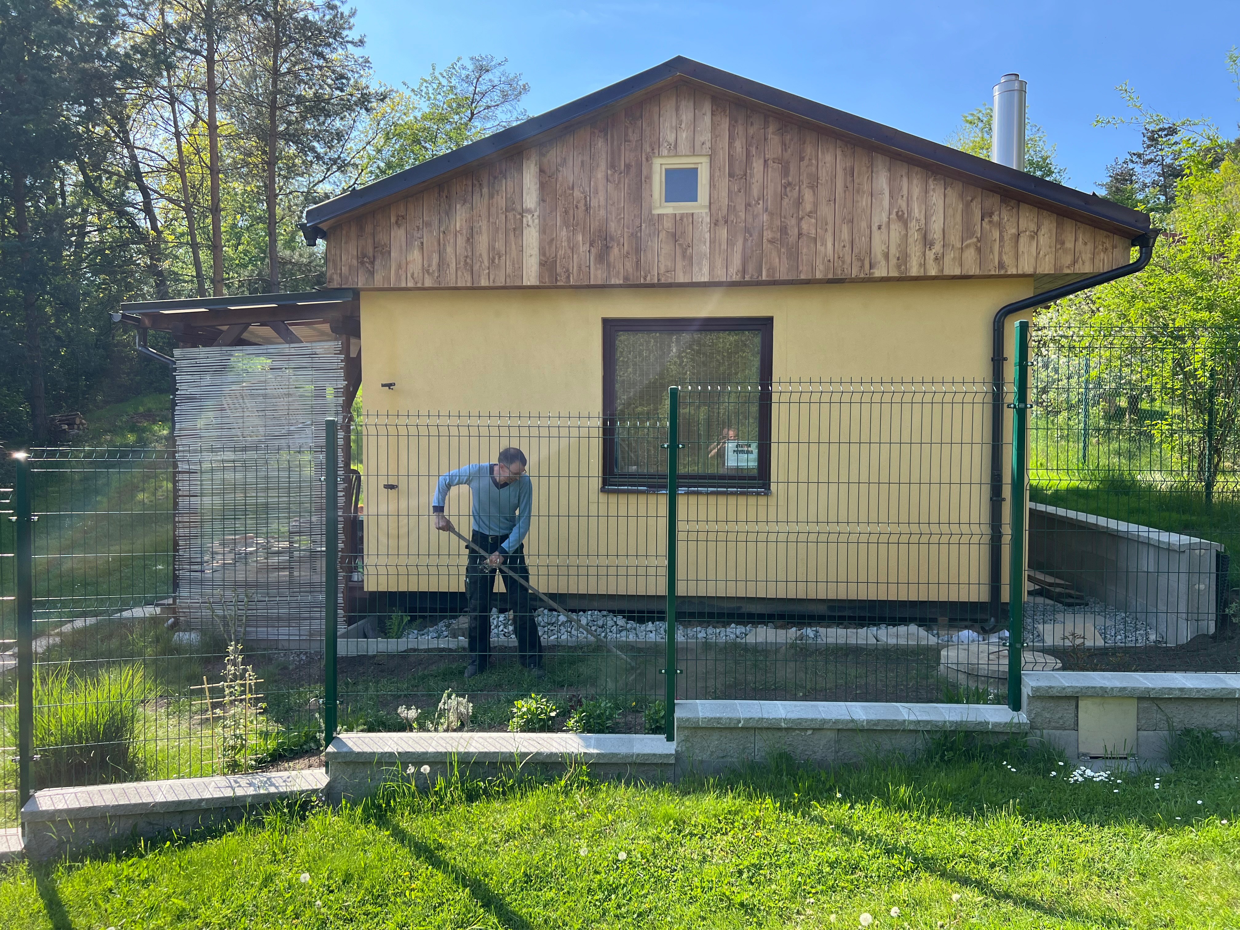 Zahradní domek s dřevěným štítem a žlutou fasádou za zeleným plotem